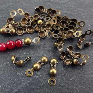 Puede incluir: Tapas de cuentas de metal de color bronce antiguo con parte superior redondeada y un bucle a cada lado. Las tapas están esparcidas sobre una superficie gris oscuro.