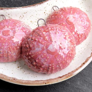 Könnte beinhalten: Drei runde, rosa dekorative Objekte mit strukturierten Oberflächen und kleinen Metallschlaufen zum Aufhängen, präsentiert auf einer gesprenkelten, cremefarbenen Keramikschale mit braunem Rand. Die Objekte haben eine glänzende Oberfläche und sind auf einer dunkelgrauen Oberfläche angeordnet.