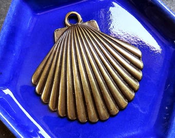 May include: A bronze-colored seashell pendant rests in a bright blue, hexagonal ceramic dish. The pendant has a loop at the top for hanging. The dish is glossy and reflects light. The background is a dark, textured surface.
