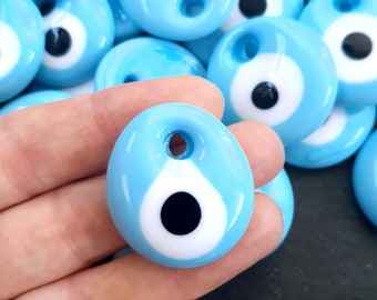 May include: A close-up of a hand holding a light blue evil eye charm with a white and black eye design. The charm has a small hole at the top for stringing.