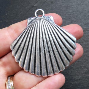 May include: A silver-coloured seashell pendant with a textured surface and a loop for hanging. The pendant has a fan-like design with detailed ridges, resembling a real seashell. The pendant is held in a hand.