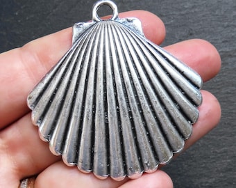 May include: A silver-colored seashell pendant with a textured surface and a loop for hanging. The pendant has a fan-like design with detailed ridges, resembling a real seashell. The pendant is held in a hand.