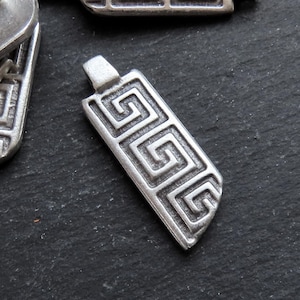 Puede incluir: Pendiente de metal plateado con un patrón geométrico de llave griega. El pendiente es rectangular y tiene un borde ligeramente redondeado.