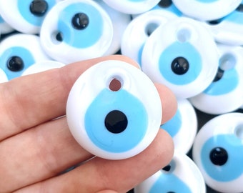 May include: A close-up of a hand holding a white and blue evil eye charm. The charm has a blue eye with a black pupil and a white ring around it. The charm is surrounded by other similar charms.