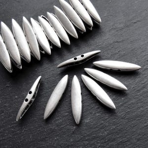 May include: A group of silver metal beads with two holes each. The beads are oval shaped and have a smooth, matte finish.