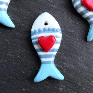 Puede incluir: Varias figuras de peces de cerámica con temática náutica. Cada pez es blanco con rayas azules, un corazón rojo y una cola azul claro. Los peces están dispuestos sobre una superficie gris oscuro.