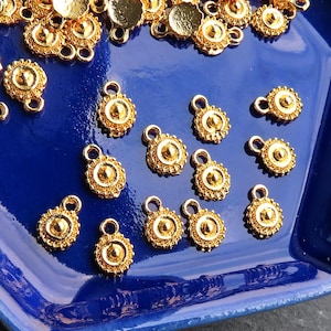 May include: Gold-colored metal charms with a circular design and small loops, scattered on a deep blue tray. The charms have a detailed, ornate pattern with a central raised circle and a decorative border.