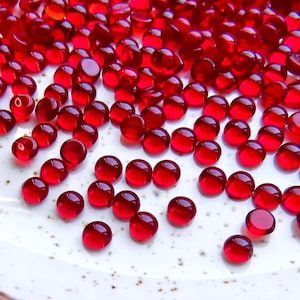 Peut inclure: Gros plan d'une assiette blanche remplie de nombreuses petites perles rondes rouges translucides. Les perles sont dispersées sur l'assiette, créant un motif visuellement attrayant. Les perles ont un aspect brillant.