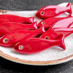 May include: A collection of small, glossy red ceramic fish-shaped beads with a single hole near the head, arranged on a white, speckled, oval-shaped ceramic dish with a brown rim.