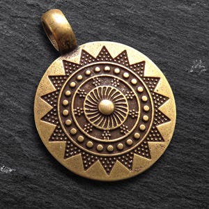 May include: A round, antique gold-colored pendant with an intricate sunburst design. The pendant features a central spiral surrounded by concentric circles and triangular patterns. A small loop is at the top for a chain.