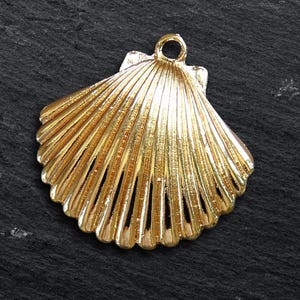 May include: A gold-colored seashell pendant with a loop for hanging. The pendant has a detailed, ridged design, resembling a scallop shell. The background is a dark, textured surface.