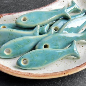 Op de afbeelding: Een verzameling turquoise keramische visbeeldjes met een gespikkelde glazuur, gerangschikt op een bord met een gespikkelde wit-bruine rand. Elke vis heeft een gat bij de kop. Decoratief item.