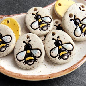 May include: A collection of ceramic bee-themed pendants and yellow disc-shaped charms arranged on a speckled, oval-shaped plate. The pendants are off-white with black and yellow bee designs. The plate has a brown rim.