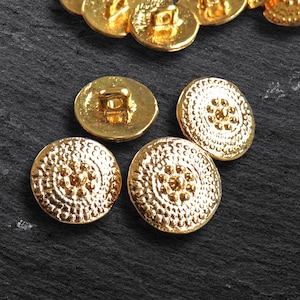 May include: Close-up of several gold-coloured buttons with a textured, circular design. The buttons have a raised, dotted pattern around a central design. Some buttons are face up, and others are face down, showing the shank.