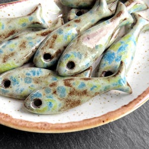 Puede incluir: Colección de cuentas de cerámica con forma de pez en tonos azules, verdes y marrones, salpicadas de color. Cada pez tiene un agujero para ensartar y se exhibe en un plato blanco con borde marrón. Suministros decorativos para manualidades.