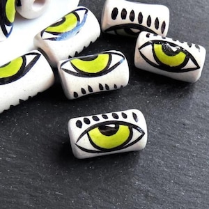 Könnte beinhalten: Weiße Keramikperlen mit schwarz und grün bemalten Augen. Die Perlen sind zylindrisch und haben eine glänzende Oberfläche.