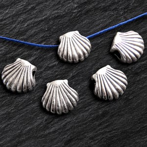May include: Five silver-colored seashell beads with a detailed fan-like design are scattered on a dark, textured surface. A blue cord runs through some of the beads, suggesting they are for jewelry making or crafts.