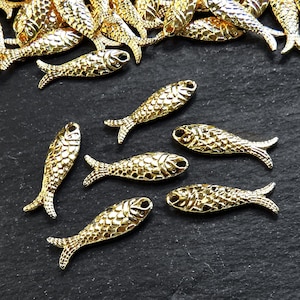 May include: A group of gold-colored fish charms, some are scattered on a black surface, while others are arranged in a row. The fish charms have a detailed, textured surface.