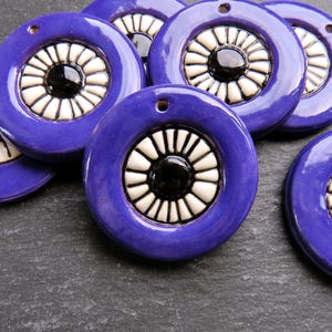 May include: Seven ceramic pendants in a deep purple hue. Each circular pendant features a white and black floral design at its centre. The pendants have a glossy finish and a small hole for threading.
