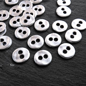 May include: A selection of silver-toned, circular buttons, each with two holes, arranged on a dark, textured background. The buttons have a slightly worn look, possibly vintage or handmade. Suitable for sewing and craft activities.