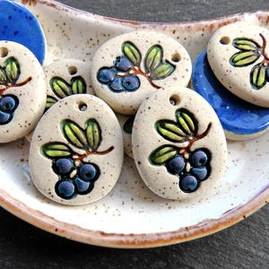 May include: Ceramic pendants with a blueberry design. The pendants are oval-shaped with a hole at the top. Each pendant features a cluster of blue blueberries with green leaves and brown stems. Two blue ceramic pieces are also visible.