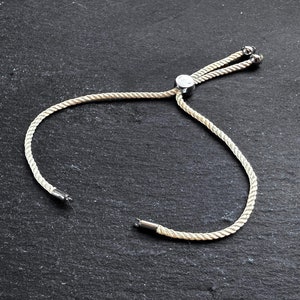Puede incluir: Un brazalete de cuerda blanca con un cierre de palanca plateado y dos cuentas plateadas en el extremo.