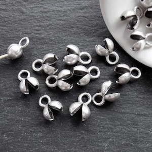 Puede incluir: Primer plano de un grupo de cuentas de crimpado de metal de color plateado. Las cuentas son pequeñas y tienen un diseño con forma de flor. Se utilizan para la elaboración de joyas.