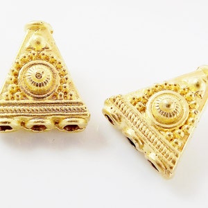 Puede incluir: Dos cuentas de metal de forma triangular de color dorado con diseños intrincados. Cada cuenta tiene un diseño circular en el centro y tres agujeros en la parte inferior.