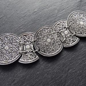 May include: Silver-toned bracelet featuring five connected, circular, ornate medallions. Each medallion displays a detailed floral and scrollwork pattern. The bracelet is linked with small metal rings, creating a flexible design. The background is a dark, textured surface.