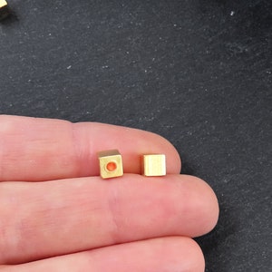 May include: Close-up of two small, gold-colored square beads. One bead has a hole through the center. The beads are on a dark surface. These beads are likely used for jewelry making or other crafts.