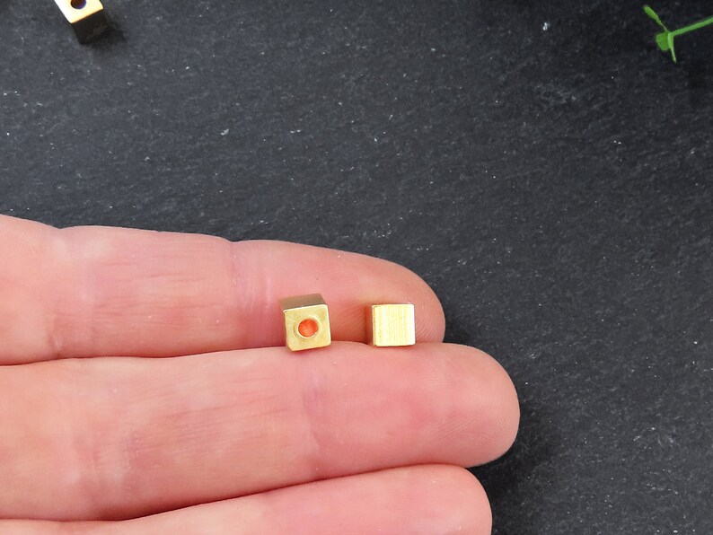 May include: Close-up of two small, gold-colored square beads. One bead has a hole through the center. The beads are on a dark surface. These beads are likely used for jewelry making or other crafts.