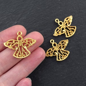 Puede incluir: Encantos de &aacute;ngel dorados con intrincados dise&ntilde;os calados. Los amuletos presentan un halo y alas extendidas, perfectos para la fabricaci&oacute;n de joyas o proyectos de artesan&iacute;a. Se ven tres amuletos, uno de ellos sostenido en una mano.