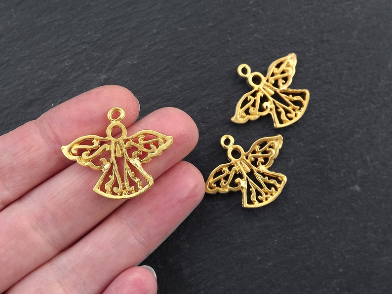 Puede incluir: Encantos de &aacute;ngel dorados con intrincados dise&ntilde;os calados. Los amuletos presentan un halo y alas extendidas, perfectos para la fabricaci&oacute;n de joyas o proyectos de artesan&iacute;a. Se ven tres amuletos, uno de ellos sostenido en una mano.