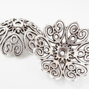 Könnte beinhalten: Zwei silberne filigranartige Metallkappen mit einem runden Loch in der Mitte. Die Kappen haben ein filigranes, kompliziertes Design mit Spiralen und Schleifen.