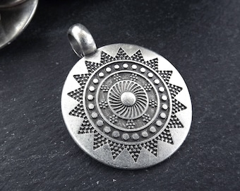 May include: Silver pendant with a circular design featuring a stylized sun with concentric circles and a starburst pattern.