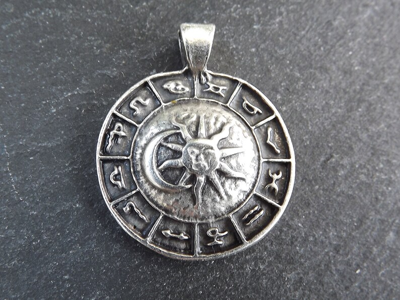 May include: Silver pendant featuring a circular zodiac wheel with the sun and moon in the center. The wheel is surrounded by the twelve zodiac signs.