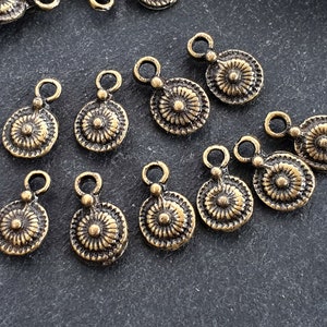May include: A collection of small, antique brass-colored charms. Each charm features a circular design with a central floral pattern and a loop at the top for attachment. The charms are scattered on a dark, textured surface.