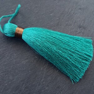 Può includere: Un singolo nappa color teal con una fascia marrone e un anello in alto. La nappa è composta da molti fili sottili, creando un aspetto morbido e fluente. Lo sfondo è una superficie scura e strutturata.
