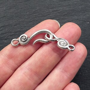 May include: A silver-coloured jewellery clasp with a hook and eye closure. The clasp has a swirling design on each end, with a curved hook in the centre. The clasp is held in a hand against a dark background.
