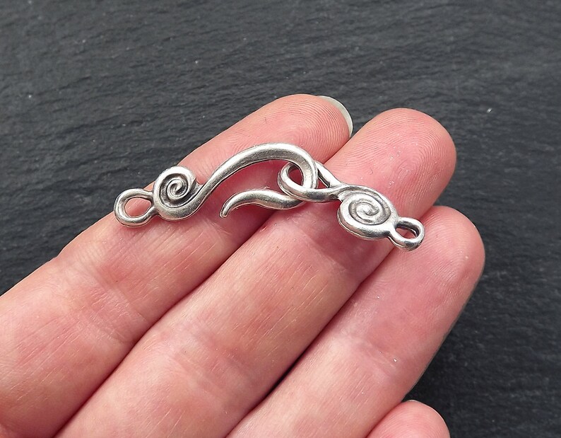 May include: A silver-coloured jewellery clasp with a hook and eye closure. The clasp has a swirling design on each end, with a curved hook in the centre. The clasp is held in a hand against a dark background.