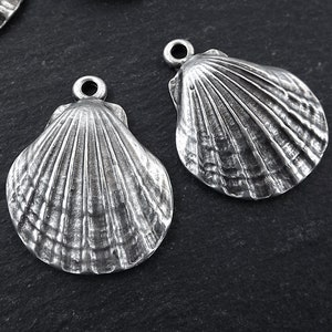 May include: Two silver-coloured seashell charms are shown in close-up. Each charm has a detailed, textured surface with radial lines and a small loop at the top for attaching to jewellery or other crafts. The charms are on a dark background.