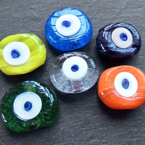 May include: Six glass evil eye charms in various colors, including blue, yellow, clear, orange, and green. Each charm features a white eye with a blue pupil.