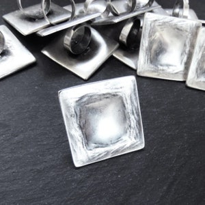 May include: A set of silver-toned metal ring holders with a square design. The holders have a raised, textured center and a smooth, flat edge.