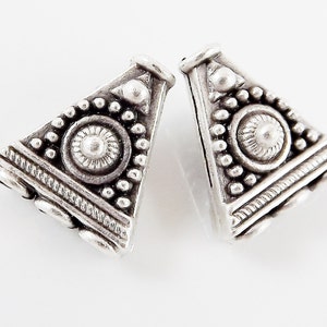 May include: Two silver triangle-shaped bead caps with intricate geometric designs. The caps have a raised centre circle with a smaller circle in the centre. The edges of the caps are decorated with small beads.