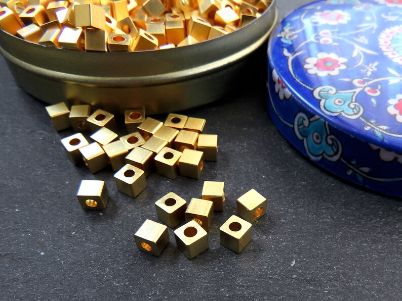 May include: A collection of small, square, gold-colored metal beads with a hole in the center. Some beads are scattered on a dark surface, while others are in a round, gold-colored tin. A blue tin with a floral design is also visible.