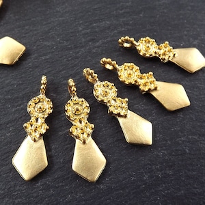 May include: Gold-colored metal jewelry findings with a diamond-shaped bottom and ornate floral-like details. The findings are arranged on a dark, textured surface. These components are likely used for jewelry making.