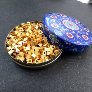 May include: A round, open tin filled with small, gold-colored, cube-shaped beads. The tin's lid, decorated with a blue floral pattern, rests beside the tin. The beads appear to be made of metal and are densely packed within the container.