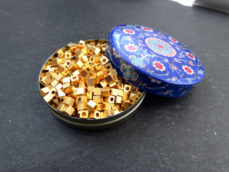 May include: A round, open tin filled with small, gold-colored, cube-shaped beads. The tin's lid, decorated with a blue floral pattern, rests beside the tin. The beads appear to be made of metal and are densely packed within the container.