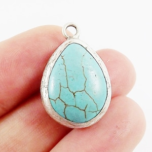 Op de afbeelding: Een druppelvormige hanger met een turquoise steen en een zilverkleurige, getextureerde rand. De steen heeft donkerbruine adering. De hanger heeft een kleine lus aan de bovenkant om op te hangen.