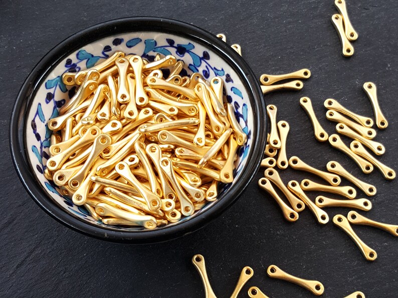 May include: A bowl filled with gold-colored metal beads. The beads are shaped like a small, elongated 'X' with a loop at each end. The bowl is white with blue and gold floral designs.
